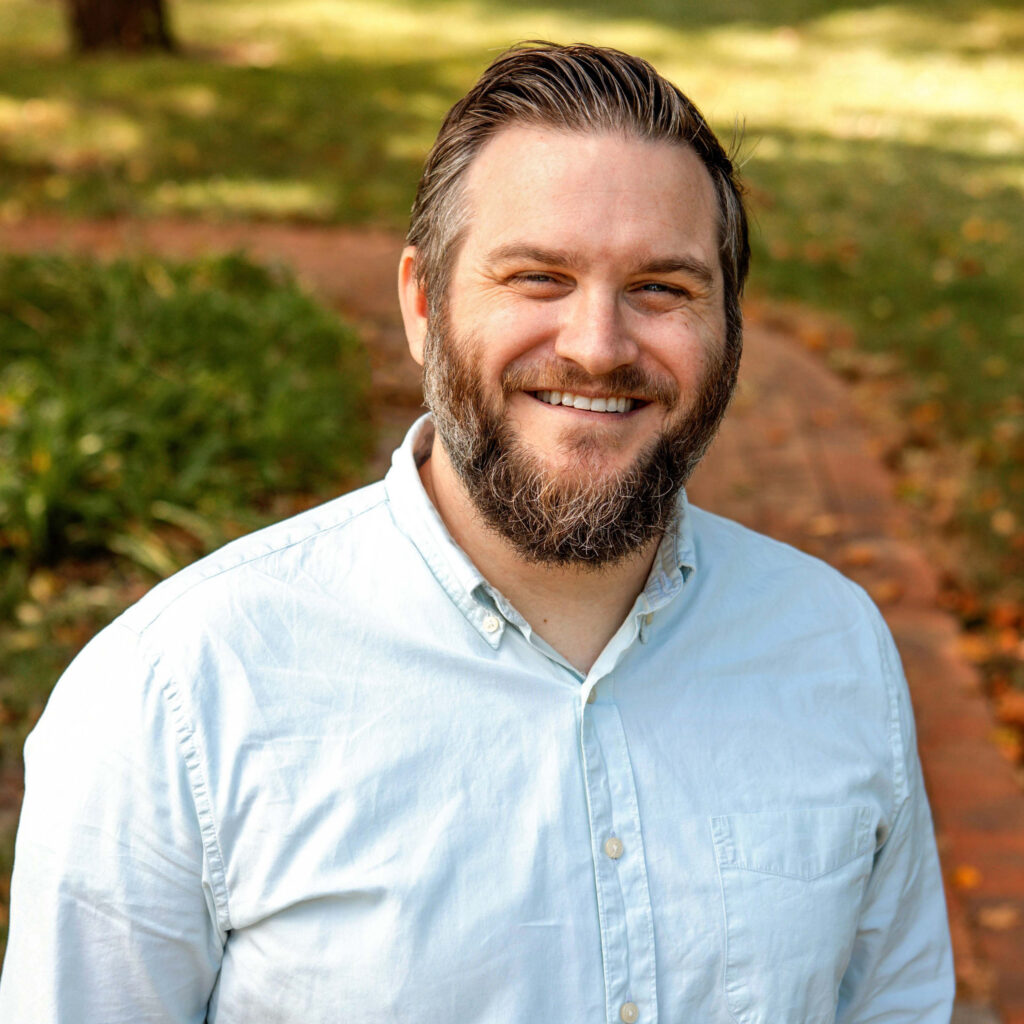 Smiling man with a neatly trimmed beard and short brown hair, wearing a light blue button-up shirt, standing on a brick path with green grass and trees in the background.