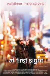 A romantic film poster featuring Val Kilmer and Mira Sorvino standing close together on a city street at night, surrounded by soft, colorful bokeh lights that suggest the bustling ambiance of New York. Kilmer wears dark sunglasses and a long coat, looking upward, while Sorvino smiles warmly at him, bundled in a winter coat. Their body language radiates connection and affection. The tagline “Inspired by a true story” hints at the heartfelt and emotional nature of the film.