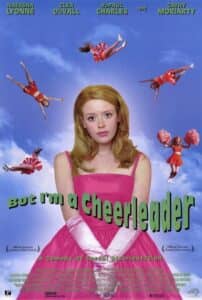 Poster for But I’m a Cheerleader, featuring Natasha Lyonne front and center in a bright pink dress and long pink gloves, looking uncertain and expressionless against a surreal blue-sky background. Around her, multiple cheerleaders in red uniforms float or tumble mid-air in exaggerated poses. The title is in bold green letters, tilted playfully: “But I’m a Cheerleader.” A tagline at the bottom reads: “A Comedy of Sexual Disorientation.” The poster also notes the film’s selection at Sundance and Toronto Film Festivals.