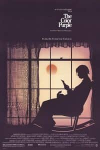A silhouetted woman sits in a chair reading a letter against the backdrop of a large window glowing with warm sunset light. The window is framed with lace curtains, and a bird perches outside on the sill. The title “The Color Purple” appears in elegant white text above, with the tagline: “It’s about life. It’s about love. It’s about us.”