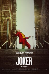 Poster for Joker, featuring Joaquin Phoenix in full clown makeup and a bright red suit, dancing triumphantly on a staircase between two brick buildings. The sky is pale and the mood is dramatic, with the film title JOKER in bold white letters below. Release date: October 4.