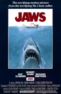 Poster for Jaws featuring a giant great white shark emerging from the deep ocean, jaws wide open, heading toward an unaware woman swimming at the surface. The tagline reads, “The terrifying motion picture from the terrifying No.1 best seller.”