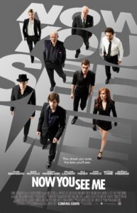 Theatrical one-sheet poster for Now You See Me set against a sleek gray background with the stylized title typography layered in large, faint white and transparent lettering behind the ensemble cast. Eight main cast members—including Jesse Eisenberg, Woody Harrelson, Isla Fisher, Dave Franco, Mark Ruffalo, Mélanie Laurent, Morgan Freeman, and Michael Caine—walk forward down a slight downward perspective dressed in sharp black suits, dress shirts, and formal wear. Just above the title at the bottom, the tagline reads, "The closer you look, the less you'll see." The film title "NOW YOU SEE ME" is printed in bold white capital letters across the lower third, above billing credits and a "COMING SOON" release notice.